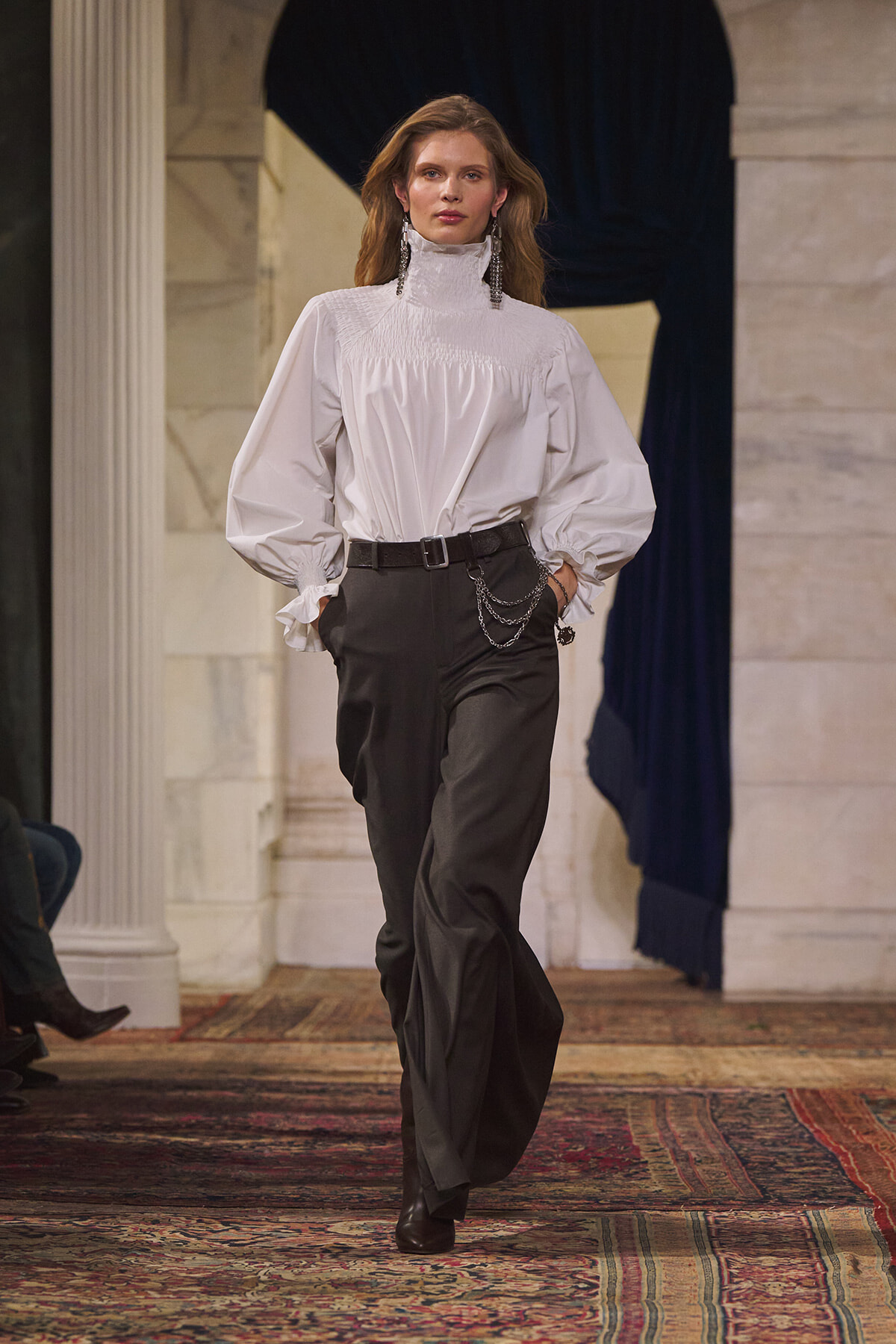 Model walking runway in a white high-neck blouse with voluminous sleeves and black wide-leg trousers, accessorized with a belt and layered chains in an elegant interior setting.