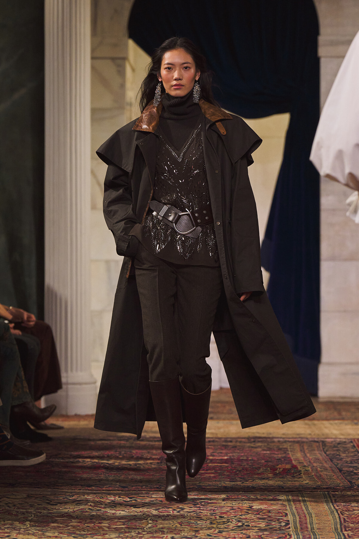 Model walks a runway in a long black trench coat over a sparkly black top, belted at the waist with knee-high boots.