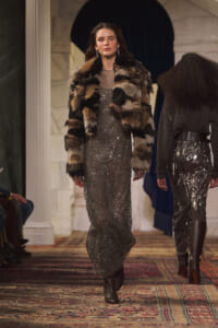Model walking a runway in a silver sequin maxi dress with a patchwork fur coat and brown boots, ornate venue backdrop behind her.