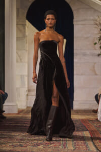 Model wearing a strapless black velvet gown with a front slit, paired with knee-high boots, walking a runway in a marble venue.