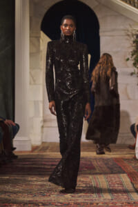Model walks a runway in a full-length black sequined turtleneck jumpsuit, striking pose under warm venue lighting.