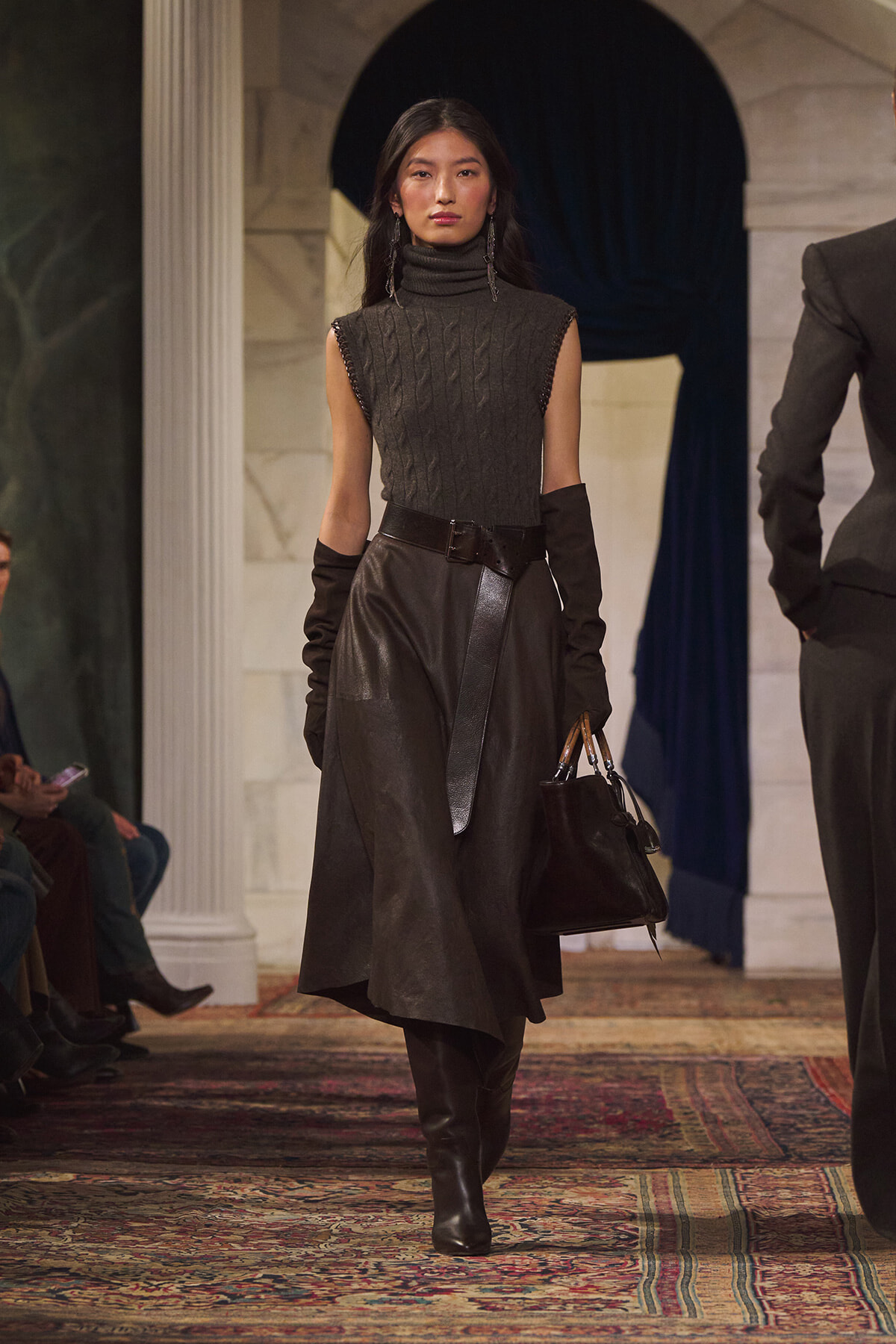 Runway model wearing a gray sleeveless turtleneck knit with a black leather MIDI skirt, belt, gloves, and knee-high boots, carrying a black handbag.