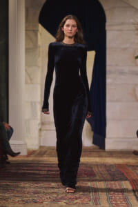 Model wearing a floor-length black velvet dress walks down a runway in a grand, marble-lined venue.