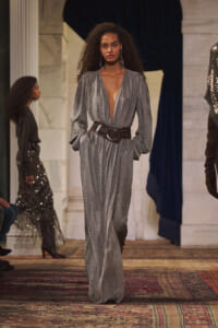 Model walks the runway in a metallic silver deep-V gown with a wide black belt, hands in pockets, on an ornate rug-lined catwalk.