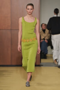 Model walking the runway in a lime-green sleeveless dress with a scalloped yellow belt and gray loafers.