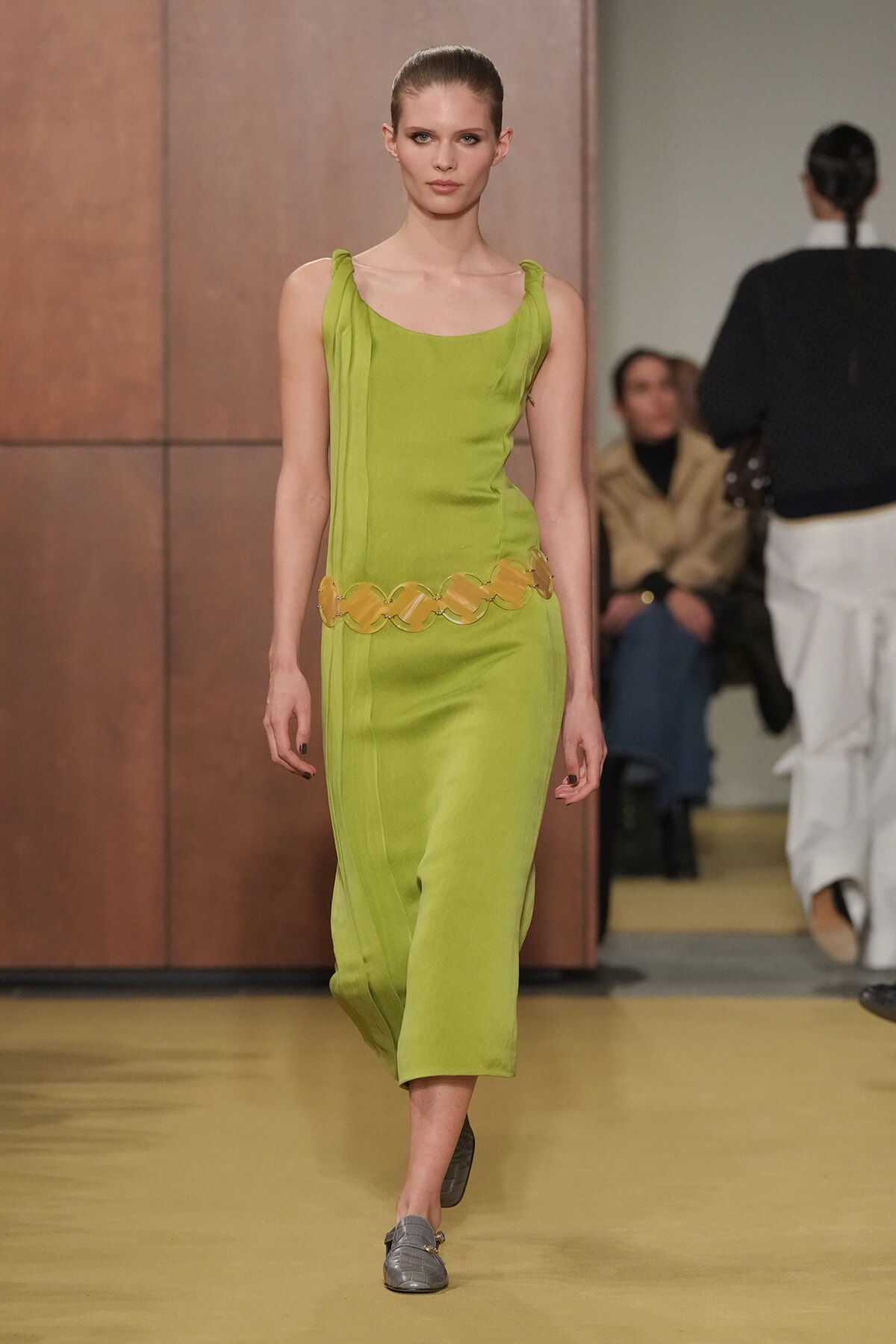 Model walking the runway in a lime-green sleeveless dress with a scalloped yellow belt and gray loafers.