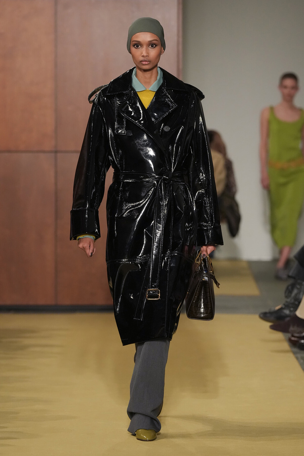 Model walks a fashion runway in a glossy black patent trench coat belted at the waist, with an olive headscarf and gray trousers, carrying a brown handbag.