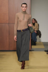 Model walking a runway in a sheer taupe turtleneck top, a long gray pleated skirt, a braided purple belt, brown boots, and a green shoulder bag.