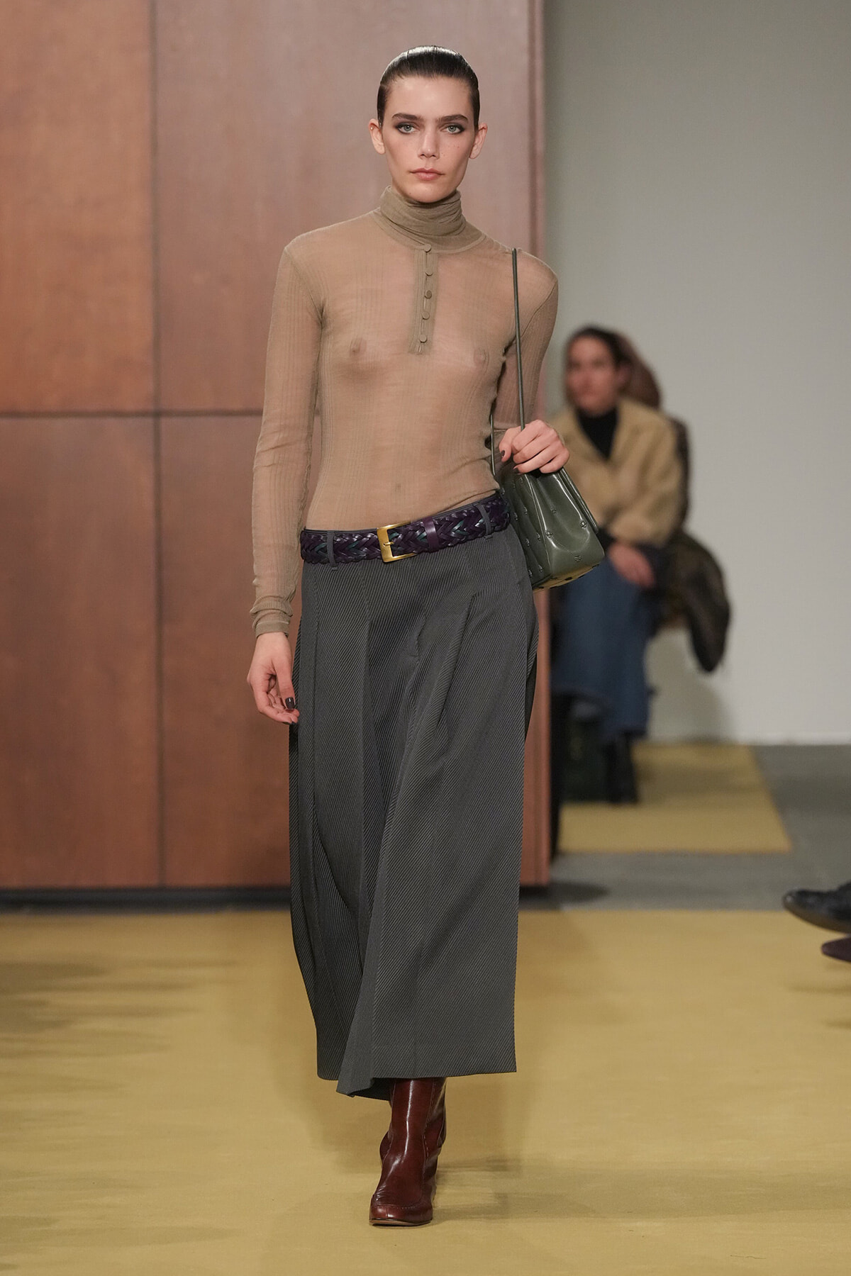 Model walking a runway in a sheer taupe turtleneck top, a long gray pleated skirt, a braided purple belt, brown boots, and a green shoulder bag.