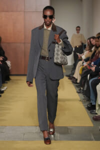 Model wearing a gray two-piece suit with a belted waist, carrying a patterned tote bag and wearing dark sunglasses on a fashion show runway.