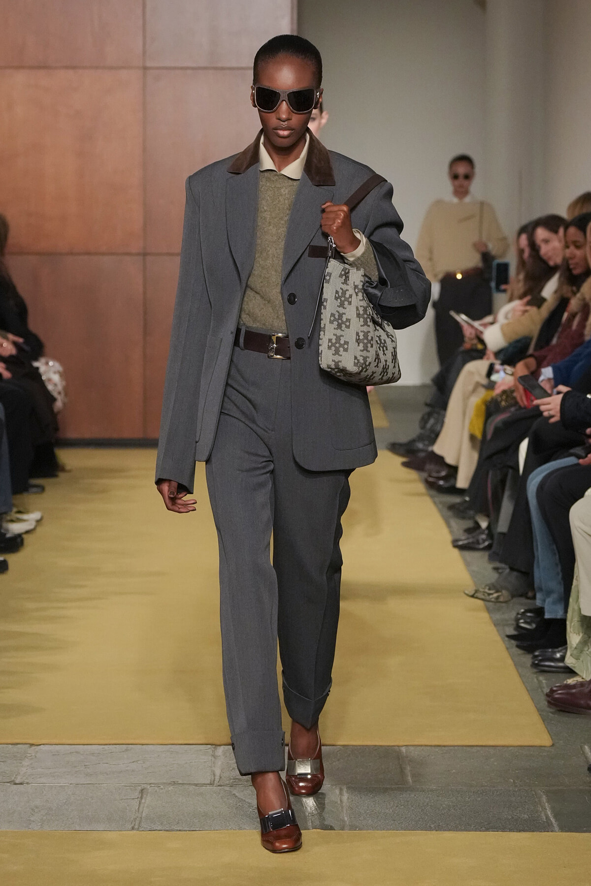 Model wearing a gray two-piece suit with a belted waist, carrying a patterned tote bag and wearing dark sunglasses on a fashion show runway.