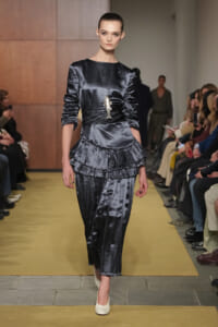 Model walks a runway in a dark gray satin dress with ruched waist and layered ruffle skirt, accessorized by a fish pendant necklace.