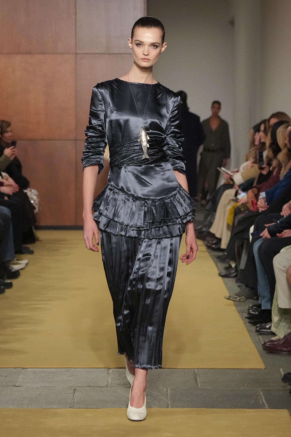 Model walks a runway in a dark gray satin dress with ruched waist and layered ruffle skirt, accessorized by a fish pendant necklace.