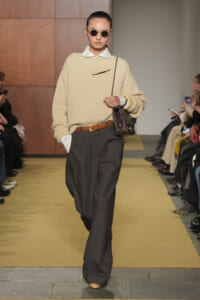 Model walks a runway in a cream textured sweater over a white collared shirt, dark wide-leg pants, a brown belt, round sunglasses, and a brown shoulder bag.