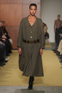 Model walks the runway in an olive-green, knee-length coat with a wide lapel, gold buttons, and a circular buckle belt, paired with black boots.