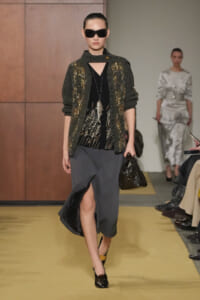 Runway model in a metallic gray cardigan over a black top with a front slit skirt, black heels, and a dark handbag with oversized sunglasses.