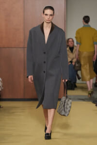 Model walking the runway in a gray oversized coat with a deep V-neck and black lapels, carrying a patterned gray handbag and wearing black heeled shoes.