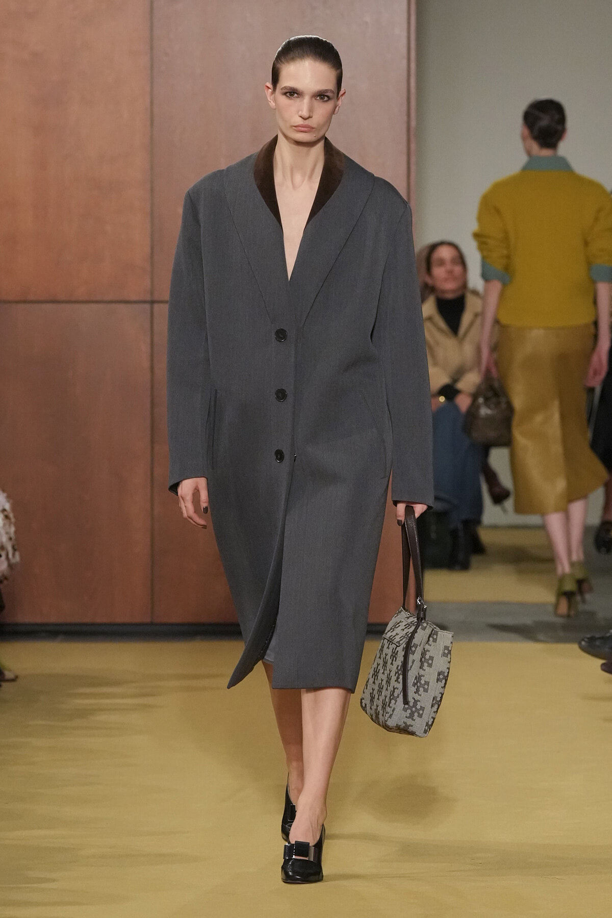 Model walking the runway in a gray oversized coat with a deep V-neck and black lapels, carrying a patterned gray handbag and wearing black heeled shoes.