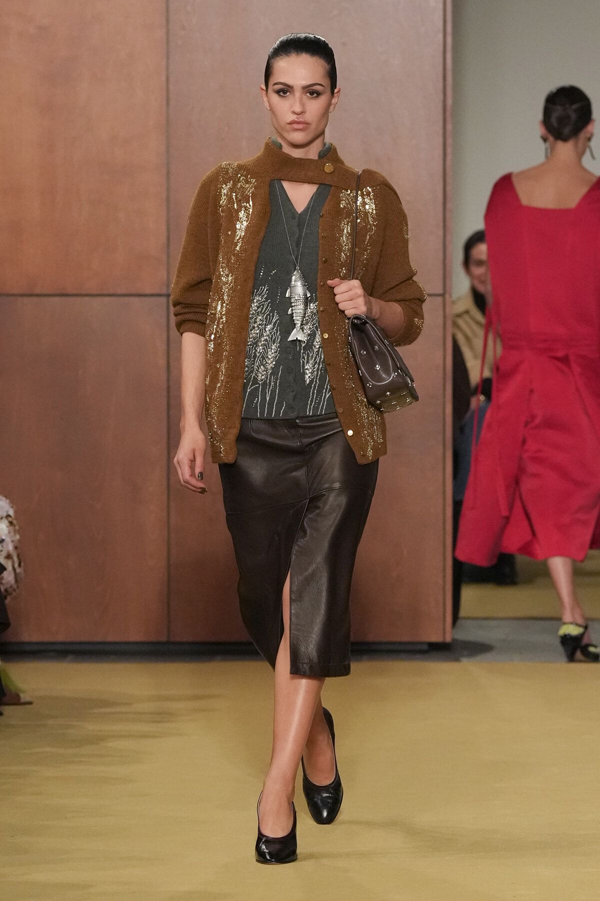 Model walking runway in a brown sequined cardigan over a dark gray top with a leaf pattern, a black leather midi skirt with a front slit, black heels, and a small black bag on a long strap.