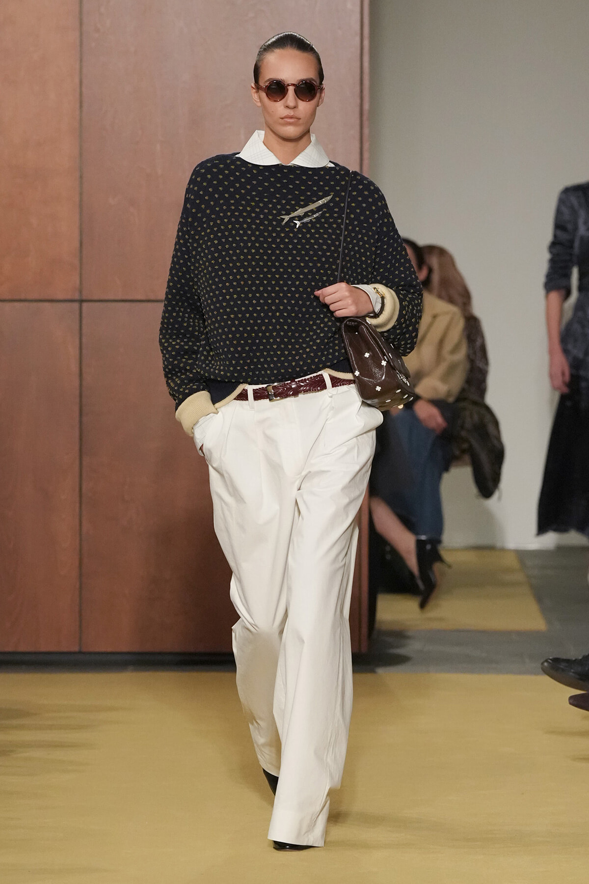 Model on a runway wearing a dark polka-dot sweater over a white collared shirt, white wide-leg pants, brown belt, and sunglasses, carrying a brown crossbody bag.