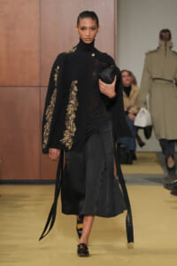 Model walks a runway in a black textured cape with gold sequin embroidery over a black turtleneck and long skirt, clutch in hand.