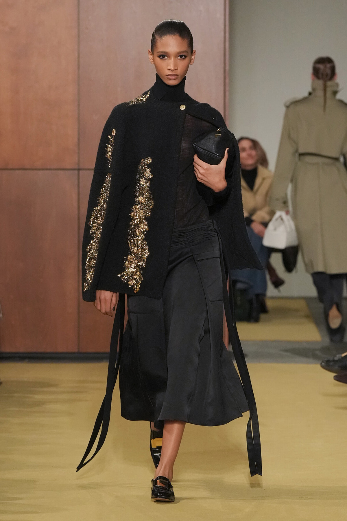 Model walks a runway in a black textured cape with gold sequin embroidery over a black turtleneck and long skirt, clutch in hand.