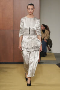 Model walks a runway in a silver satin ensemble with a ruched waist and multi-layered ruffle peplum.