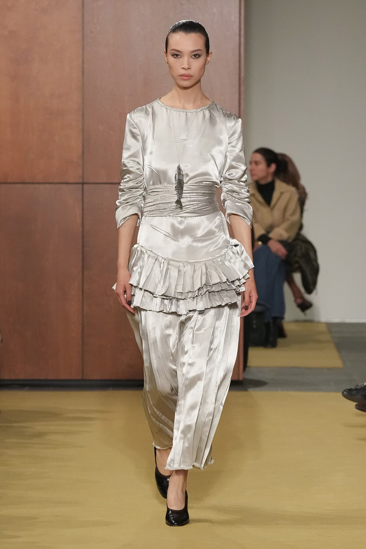 Model walks a runway in a silver satin ensemble with a ruched waist and multi-layered ruffle peplum.