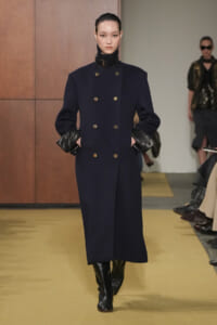 Model on a runway wearing a navy double-breasted wool coat with gold buttons, black leather gloves, and knee-high boots.
