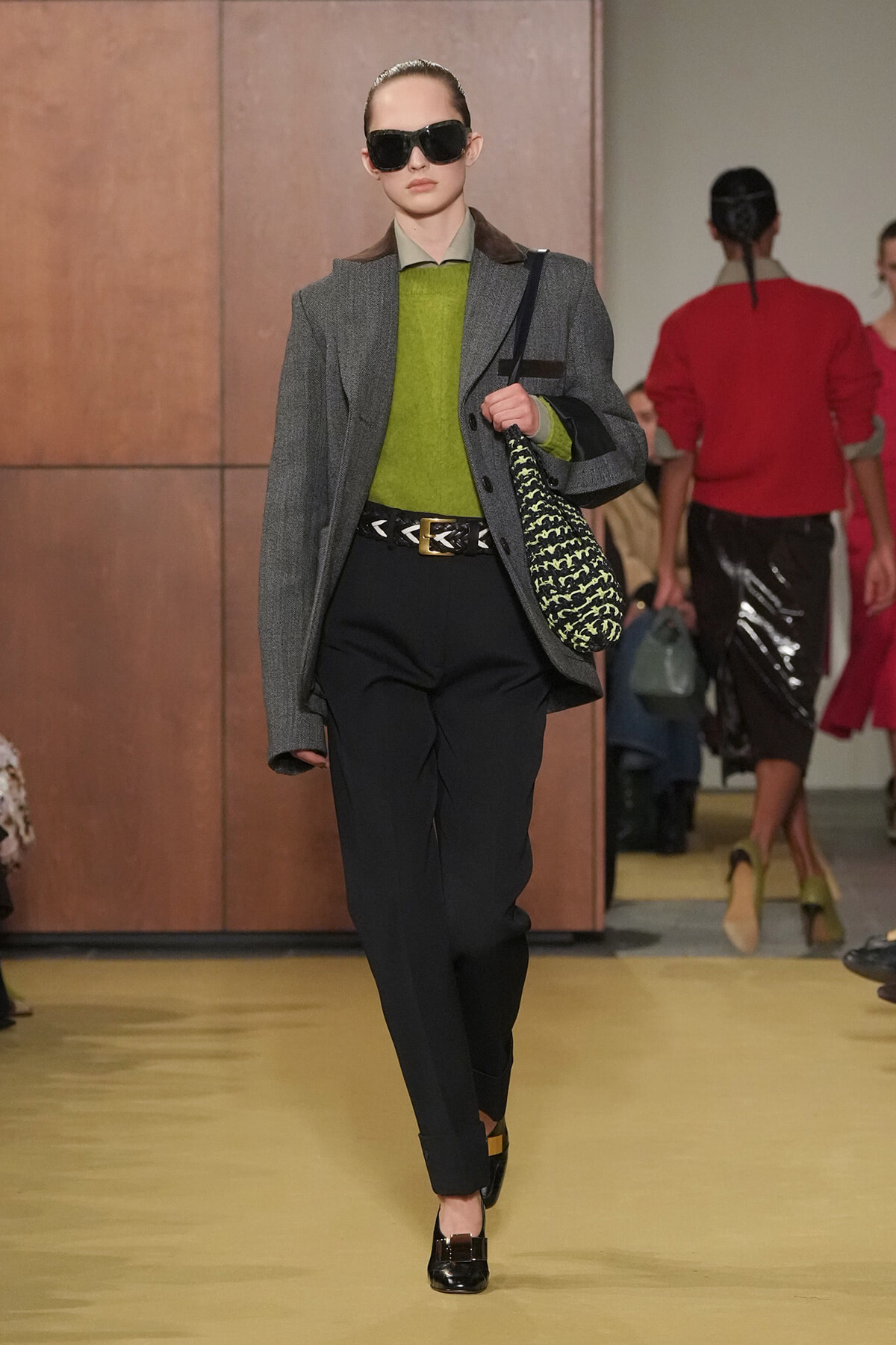Model on runway in a gray blazer over a lime sweater, black trousers, belt, oversized sunglasses, carrying a patterned green-and-black handbag.