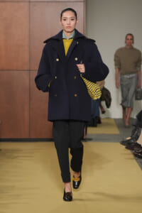 Runway model in a navy double-breasted coat with gold buttons, mustard sweater, dark tapered pants, and a yellow-black woven bag over her shoulder.