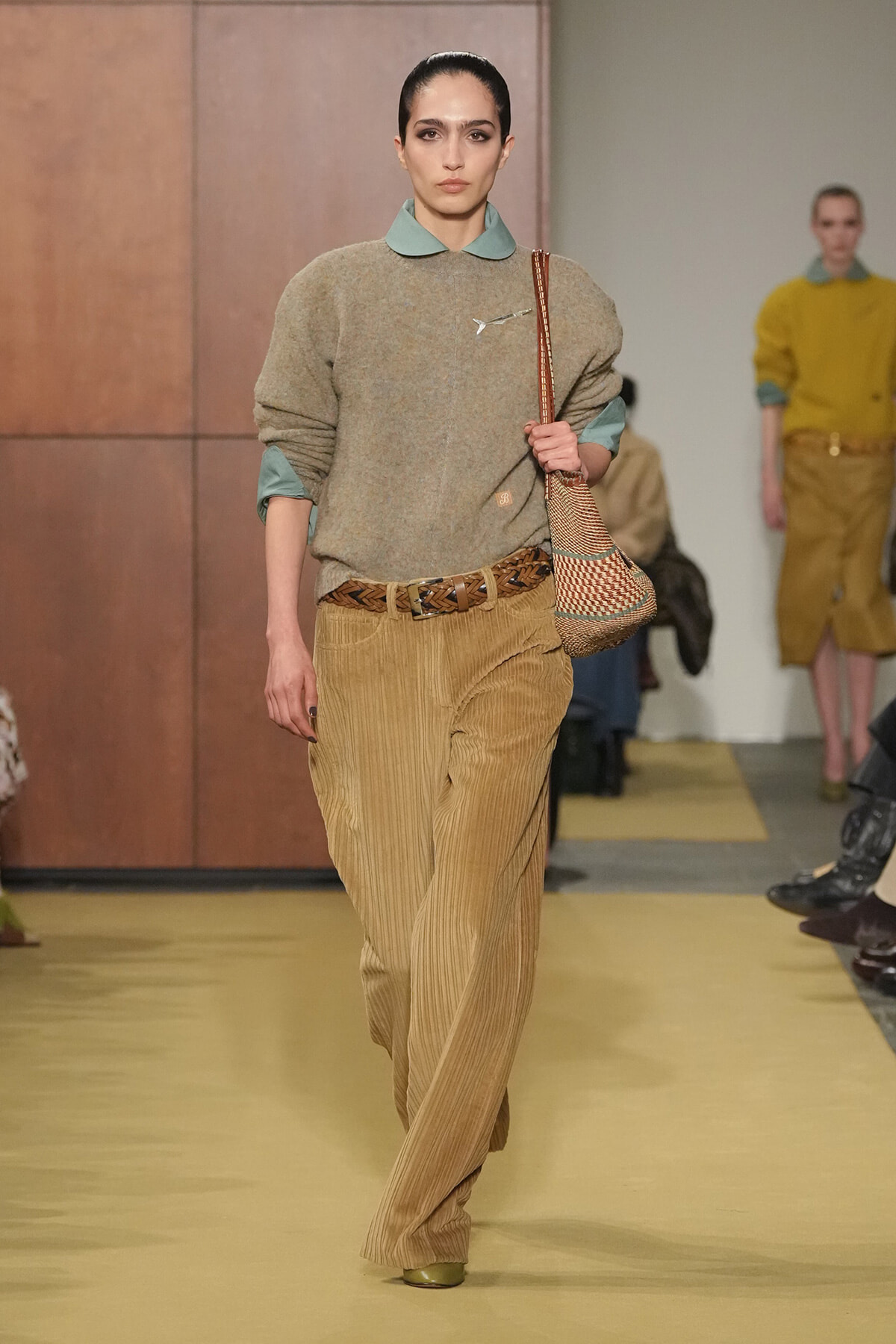 Model walking a runway in a beige knit sweater with a teal collar, paired with tan corduroy wide-leg pants and a braided belt, carrying a woven bag on her shoulder.