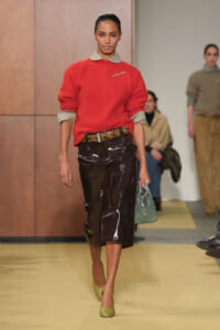 Model walking the runway in a red knit sweater with rolled gray cuffs, a light gray collar, and a brown patent leather midi skirt paired with a braided belt; light green flats and a sage handbag.