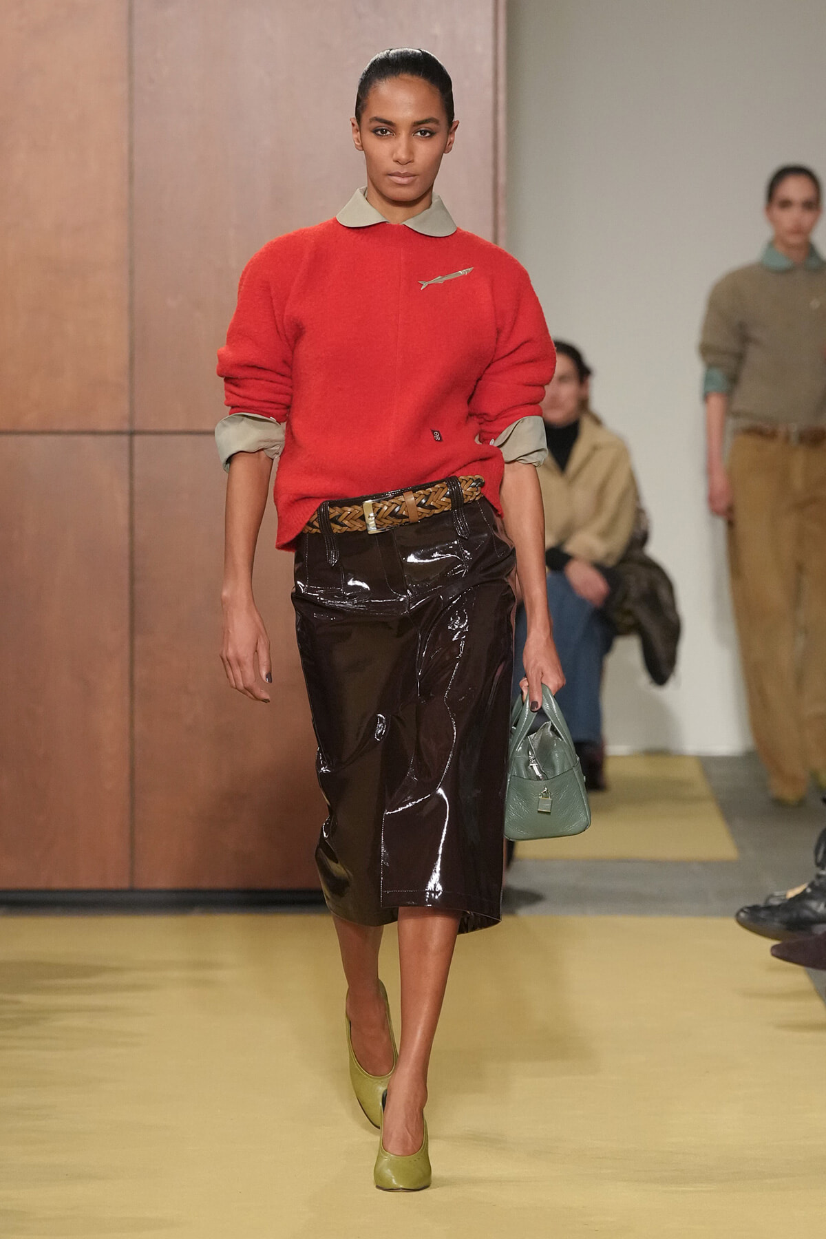 Model walking the runway in a red knit sweater with rolled gray cuffs, a light gray collar, and a brown patent leather midi skirt paired with a braided belt; light green flats and a sage handbag.