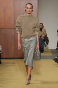 Model walking runway in a beige fuzzy oversized sweater with rolled sleeves and a sage satin midi skirt, paired with a braided belt and gray handbag.