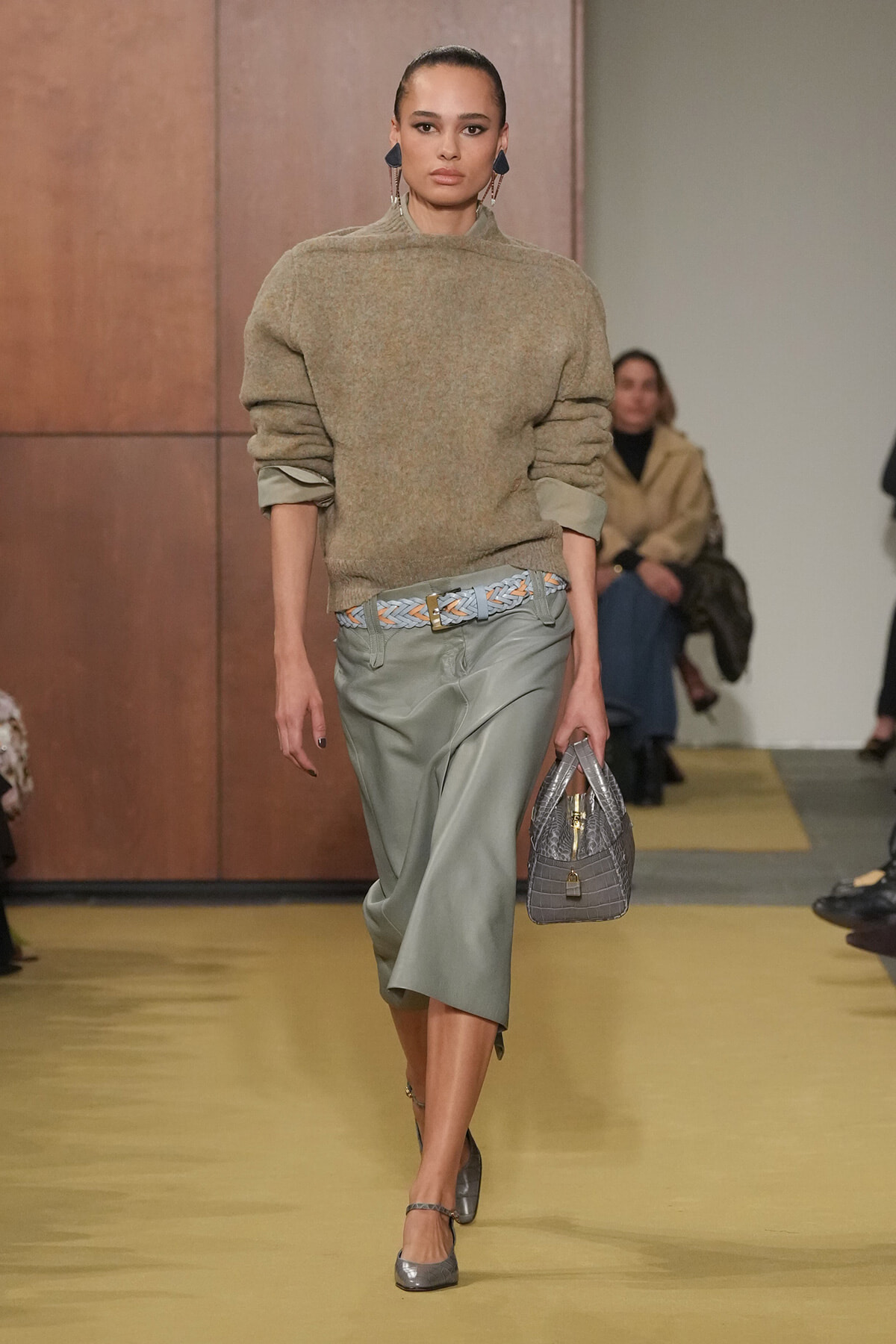 Model walking runway in a beige fuzzy oversized sweater with rolled sleeves and a sage satin midi skirt, paired with a braided belt and gray handbag.