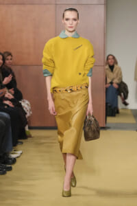 Model on a runway wearing a mustard yellow sweater with a green collar, a mustard leather midi skirt, a braided belt, greenish shoes, and a brown handbag.