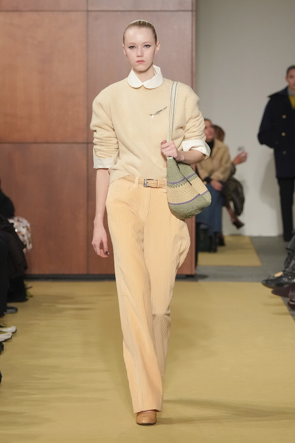 Model walks a runway in a beige knit sweater with a white collar, pale striped wide-leg pants, and a woven shoulder bag.