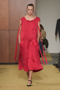Model walks a red midi dress with cape-like sleeves and long fabric ties, on a fashion runway with a neutral backdrop.