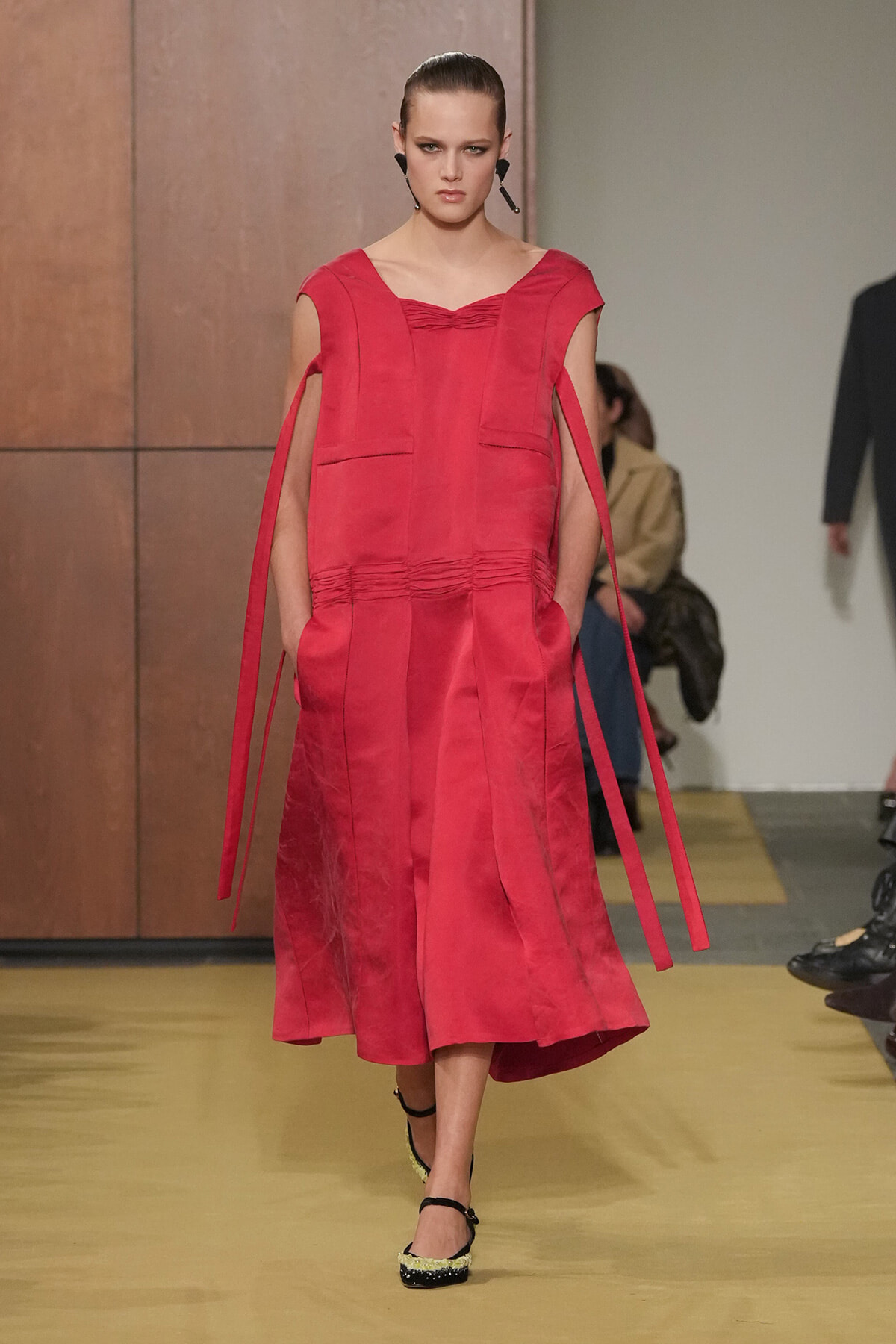 Model walks a red midi dress with cape-like sleeves and long fabric ties, on a fashion runway with a neutral backdrop.