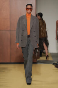 Model on a runway wearing an oversized grey blazer over an orange turtleneck and matching grey trousers, carrying a brown handbag and wearing orange sunglasses.