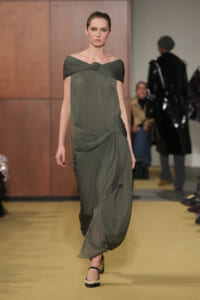Model walking a runway in an olive-green draped gown with a twisted neckline and cinched waist.