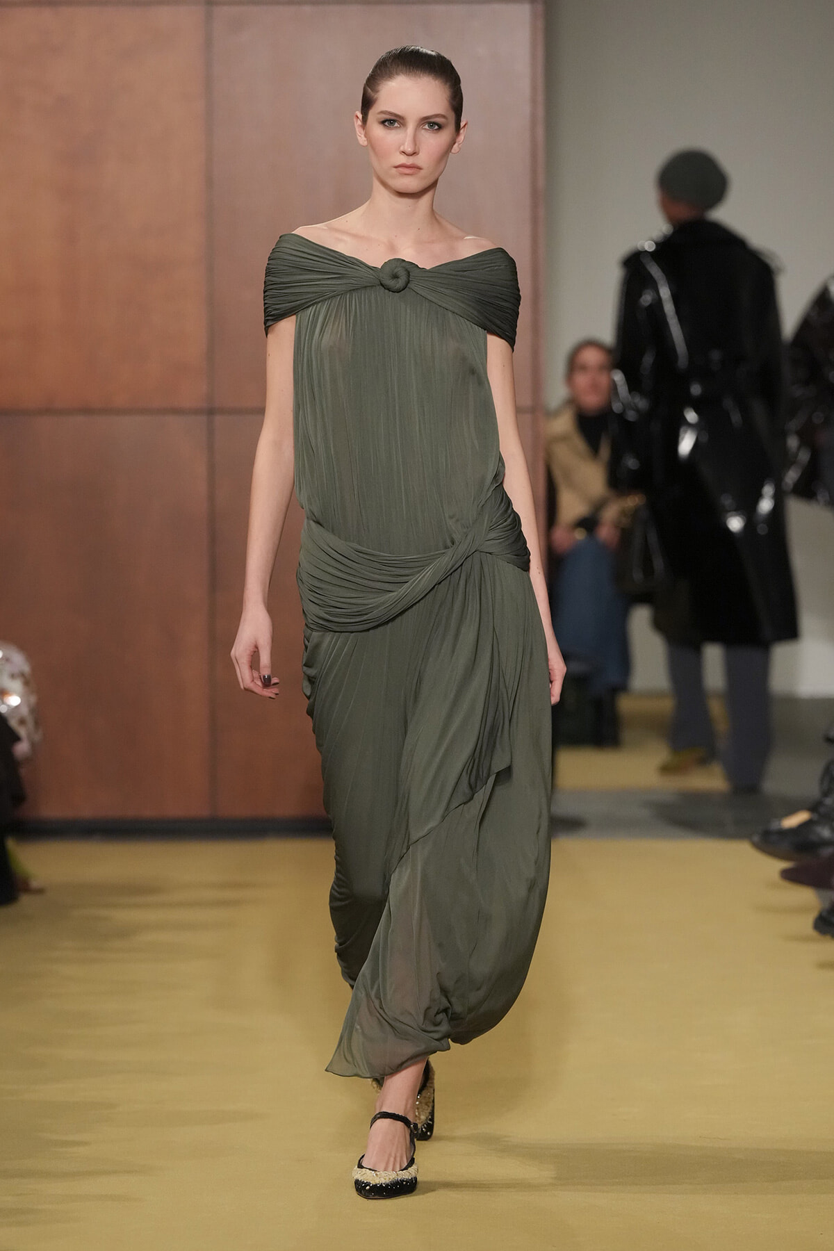 Model walking a runway in an olive-green draped gown with a twisted neckline and cinched waist.