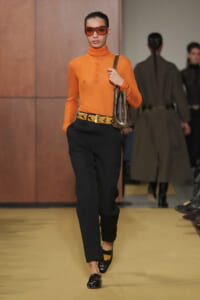 Model walking a runway in an orange turtleneck top, black trousers with a braided yellow belt, a brown shoulder bag, and orange sunglasses.