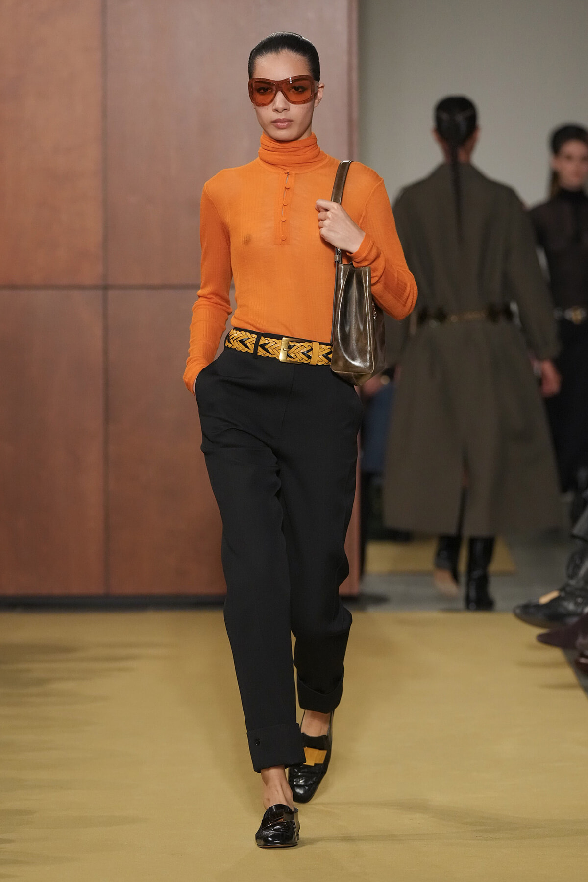 Model walking a runway in an orange turtleneck top, black trousers with a braided yellow belt, a brown shoulder bag, and orange sunglasses.