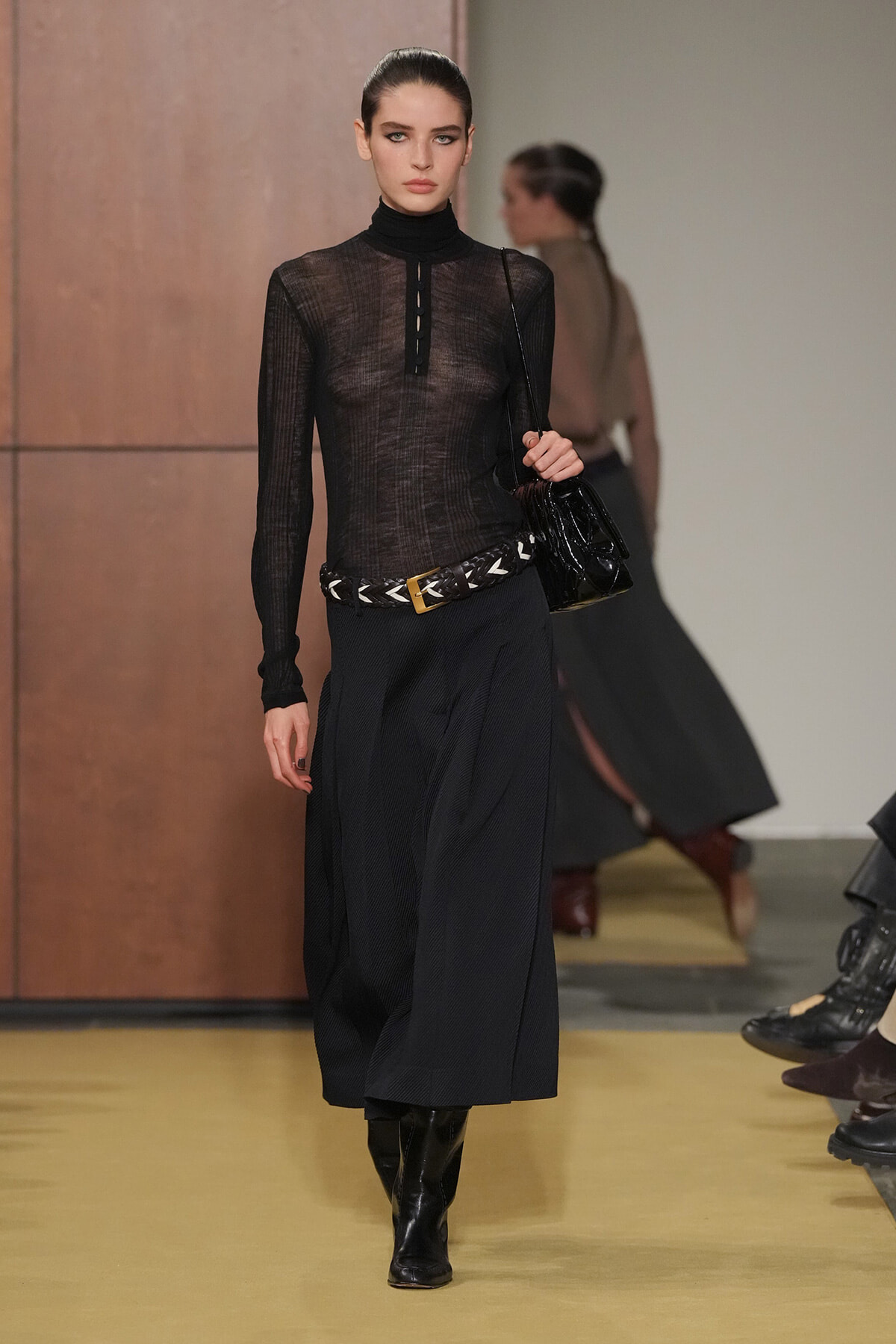 Model walks the runway in a sheer black turtleneck top, a long dark skirt with a braided belt, black boots, and a black handbag slung over her shoulder.