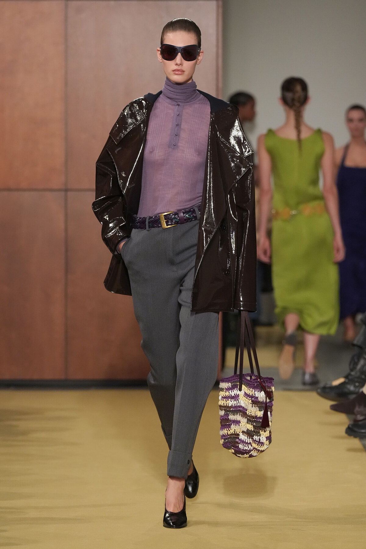 Model walking runway in a glossy dark coat over a lavender turtleneck, gray trousers, and black heels, holding a purple‑ivory woven bag and wearing oversized sunglasses.