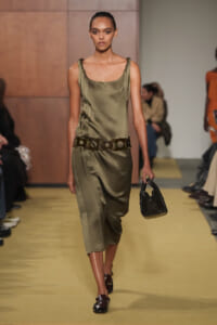 Model walking a fashion runway in an olive satin sleeveless dress with a wide belt and a small black crocodile-textured handbag.