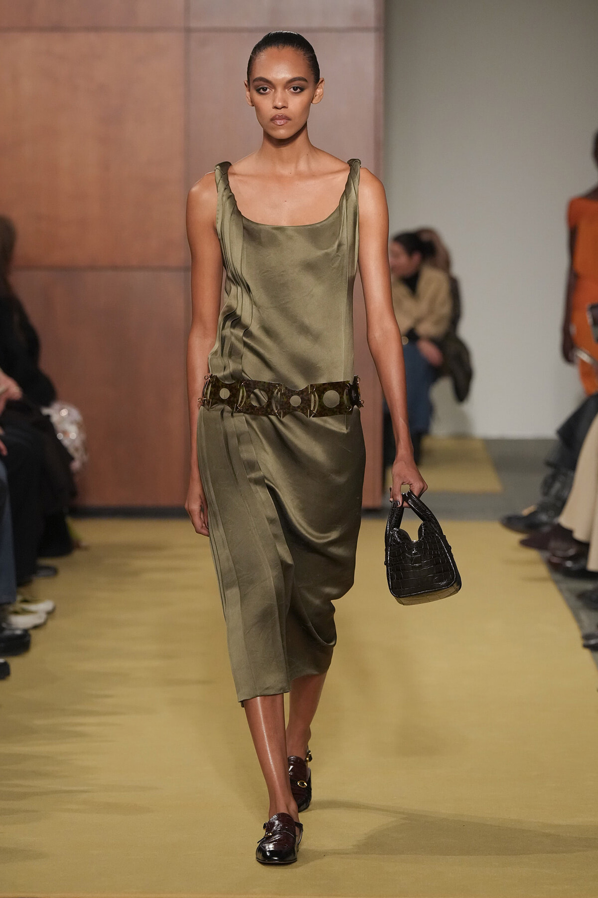 Model walking a fashion runway in an olive satin sleeveless dress with a wide belt and a small black crocodile-textured handbag.
