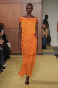 Model wearing an orange off-shoulder draped gown with a front knot walking a runway.